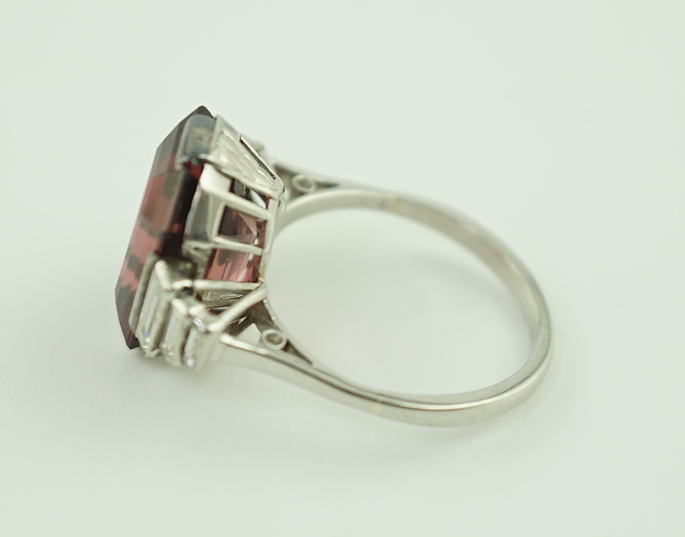 An Art Deco platinum and single stone emerald cut deep pink tourmaline set dress ring, with graduated six stone baguette cut diamond set shoulders